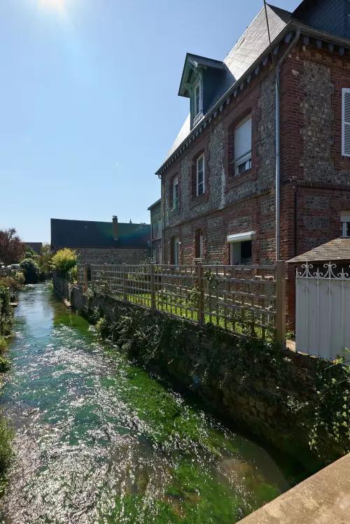 Ein Haus steht quasi direkt am Fluss. Auf der Flussoberfläche spiegeln sich die Sonnenstrahlen, die ganze Szene ist etwas sehr grell und lichtüberflutet.