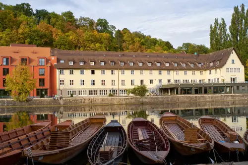 Blick auf die Jugendherberge Tübingen, mit Altbau und Anbau und mit deren heller und roter Fassade, im Frühherbst, vom Steinlachhafen aus, an dessem Steg einige Stocherkähne vertäut sind.