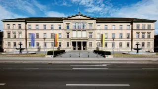 Frontalblick auf die "Neue Aula" - mit den Flaggen der EU, BRD und Baden-Württemberg - der Universität Tübingen an einem sonnigen Tag mit blauem Himmel.