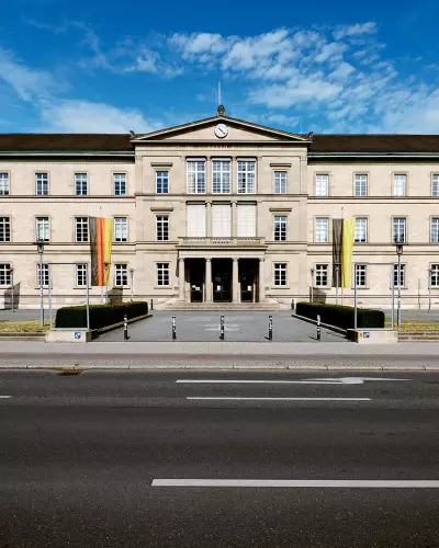 Frontalblick auf die "Neue Aula" - mit den Flaggen der EU, BRD und Baden-Württemberg - der Universität Tübingen an einem sonnigen Tag mit blauem Himmel.