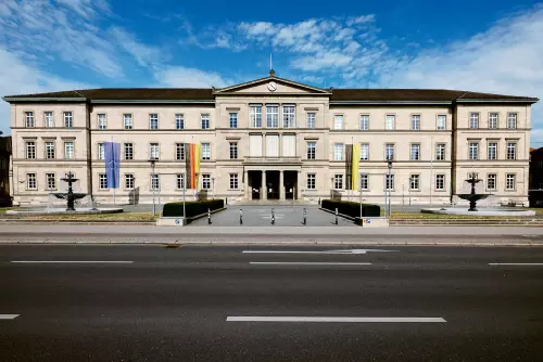 Frontalblick auf die "Neue Aula" - mit den Flaggen der EU, BRD und Baden-Württemberg - der Universität Tübingen an einem sonnigen Tag mit blauem Himmel.