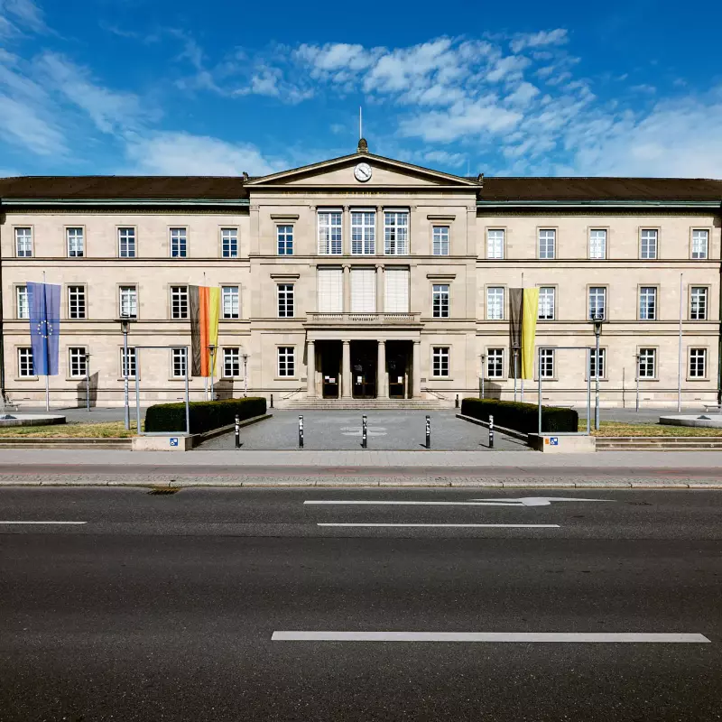 Frontalblick auf die "Neue Aula" - mit den Flaggen der EU, BRD und Baden-Württemberg - der Universität Tübingen an einem sonnigen Tag mit blauem Himmel.
