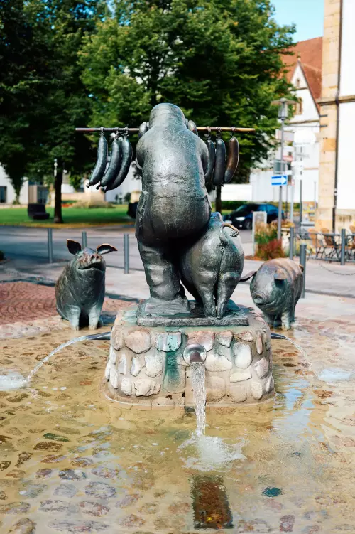 Der "Wurstträgerbrunnen" auf dem Marktplatz von Versmold. Ein Mann trägt sechs Wurstpaare auf einer Stange, während sich vor und neben ihm drei lebendige Schweine tummeln.