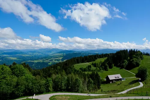 Blick von der Aussichtsplattform auf dem Pfänder, bei Bregenz, Richtung Südosten auf Täler, Bäume, Wanderweg und Alpenpanorama an einem sonnigen Tag, mit ein paar weißen Wolken vor blauem Himmel.