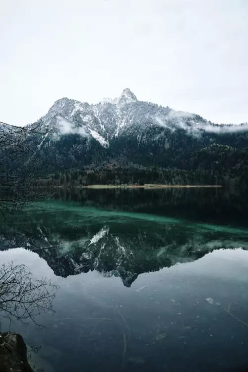 Blick vom Rundwanderweg Alpsee auf den am Gipfel leicht verschneiten Gassenthomaskopf, der sich in der grünlichen, ganz ruhigen Seeoberfläche spiegelt.