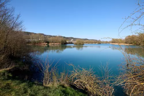 Blick vom kurzen Rundwanderweg um den Hirschauer Baggersee auf den See, das Beton- und Kieswerk im Hintergrund, an einem sonnigen Apriltag.