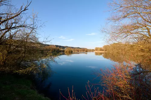 Blick vom kurzen Rundwanderweg, zweite Runde bei anderem Licht, um den Hirschauer Baggersee auf den See, das Beton- und Kieswerk im Hintergrund, an einem sonnigen Apriltag.