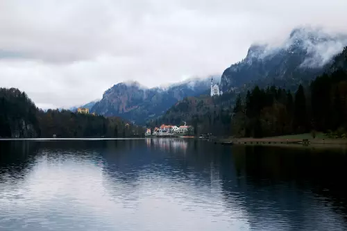 Entfernter Blick vom Rundwanderweg Alpsee auf das Schloss Neuschwanstein und seine touritischen Anlagen im Tal an einem bewölkten Novembertag.