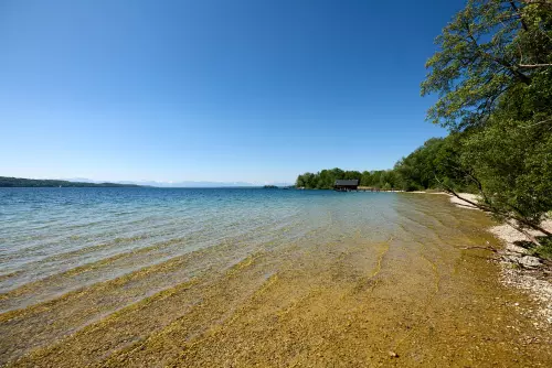 Blick vom Strand des Ammersees, mit plätschernden, kleinen Wellen an einem sonnigen Junitag mit wolkenfreiem, blauem Himmel.