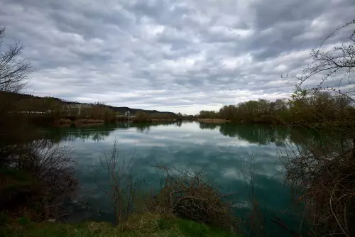 Blick vom kurzen Rundwanderweg, dritte Runde, an einem anderen Tag, um den Hirschauer Baggersee auf den See, das Beton- und Kieswerk im Hintergrund, an einem sonnigen Apriltag.