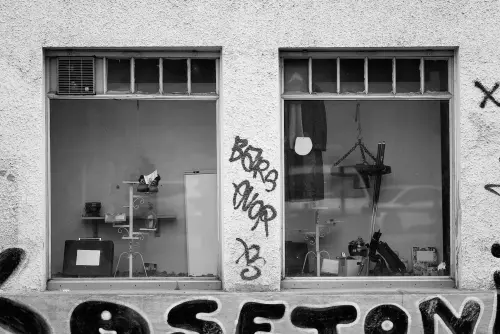 SW-Aufnahme zweier Schaufenster, mit skurrilen Dekoelementen, die von einer groben Fassadenoberfläche mit einigen gerade noch erkennbaren Graffiti-Tags eingerahmt sind.