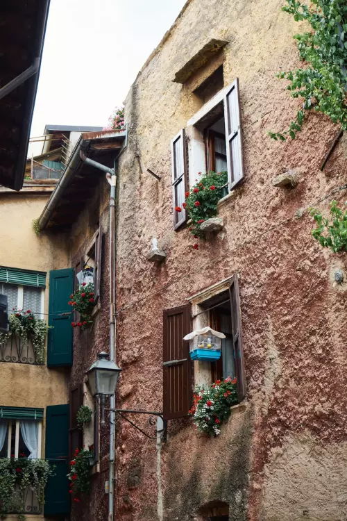 Seitengasse in Torri del Benaco; Die ansonsten beschauliche Kulisse mit rustikalen Fassaden, rot blühenden Geranien, grünen Fensterläden... wird durch einen auffällig blauen und viel zu kleinen Vogelkäfig inklusive „Insasse“ getrübt.
