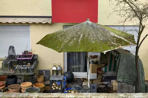 Vollgestellte Terrasse, auf der zahlreiche leere Blumentöpfe und Laternen gelagert werden plus ein maroder Strandkorb und ein aufgespannter Sonnenschirm, der komplett von Moos überwachsen ist.