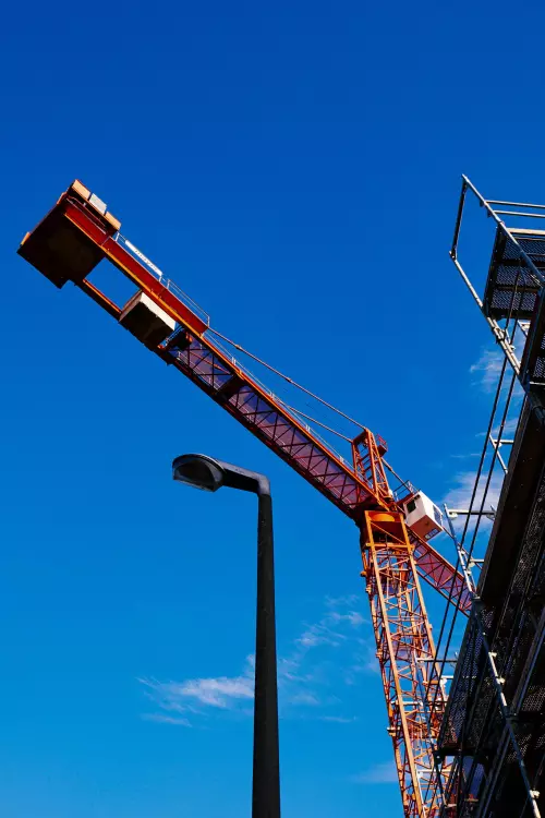 Komplett orangefarbener Baukran vor blauem Himmel. Im Vordergrund eine Straßenlaterne und rechts im Bild sind Baugerüste zu sehen.