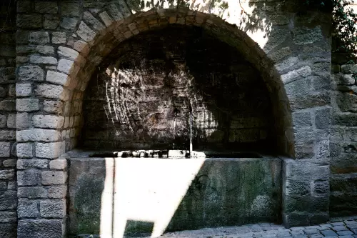 Historischer Quellbrunnen, eingelassen in der aus groben Sandsteinen gefertigten Stützmauer der Neckarhalde in Tübingen, wobei die Sonnenstrahlen, von der Wasseroberfläche reflektiert, den dunklen Mauerhintergund erleuchten.