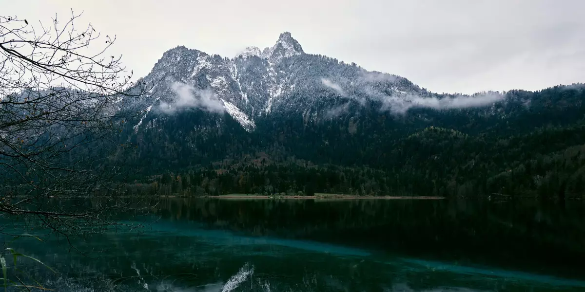 Blick vom Rundwanderweg Alpsee auf den am Gipfel leicht verschneiten Gassenthomaskopf, der sich in der grünlichen, ganz ruhigen Seeoberfläche spiegelt.