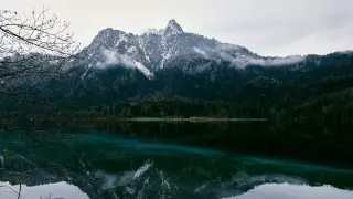 Blick vom Rundwanderweg Alpsee auf den am Gipfel leicht verschneiten Gassenthomaskopf, der sich in der grünlichen, ganz ruhigen Seeoberfläche spiegelt.